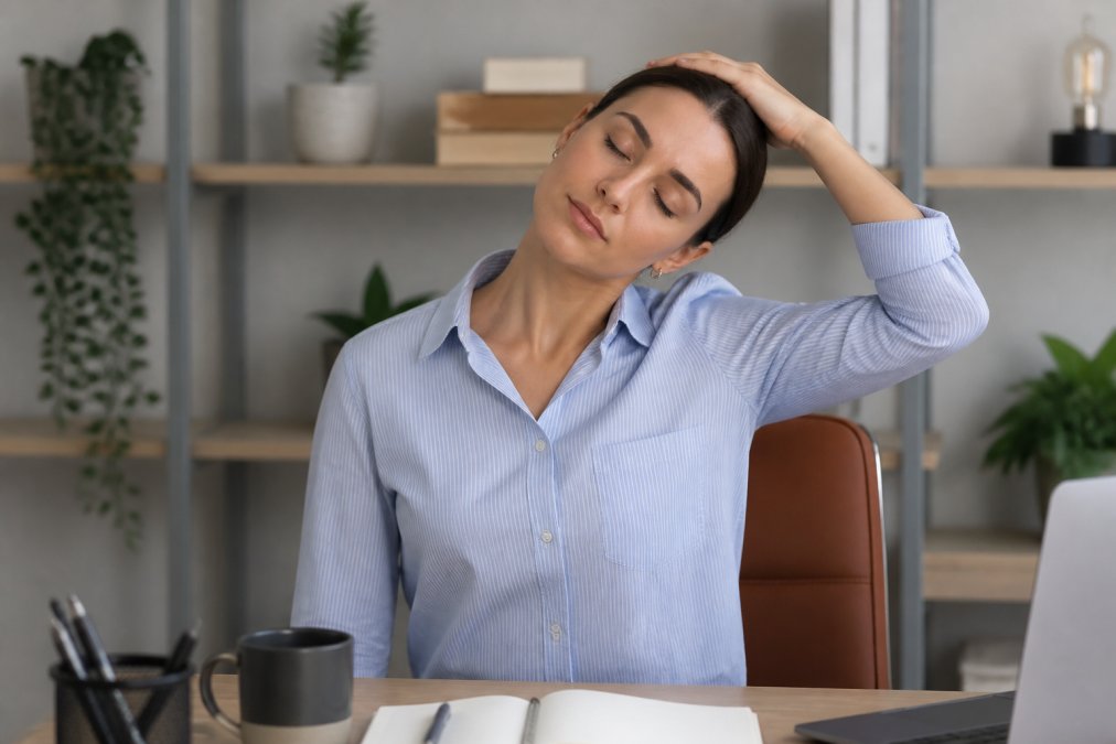 Woman performing gentle neck stretch at desk, tilting head toward shoulder with proper posture, natural office lighting showing correct form for chronic neck pain relief exercise