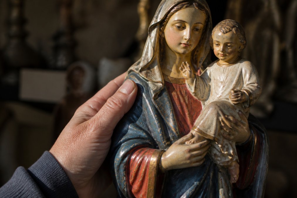 Close-up van een hand die voorzichtig een antiek gipsen Mariabeeld met kindje Jezus vasthoudt, warm invallend zijlicht accentueert de handbeschilderde details en de natuurlijke craquelé in de verflaag