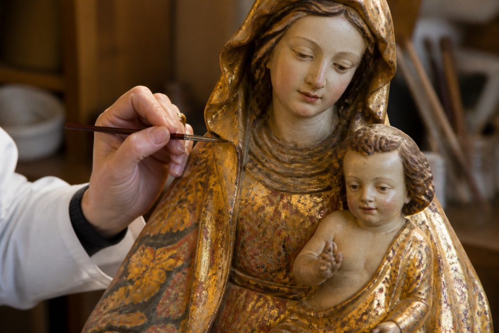 Handen van een restaurateur die met een fijn kolinsky-penseel zorgvuldig kleur aanbrengt op een antiek polychroom religieus beeld, zichtbaar detail van het vergulde oppervlak en de pigmenten in warm atelierlicht