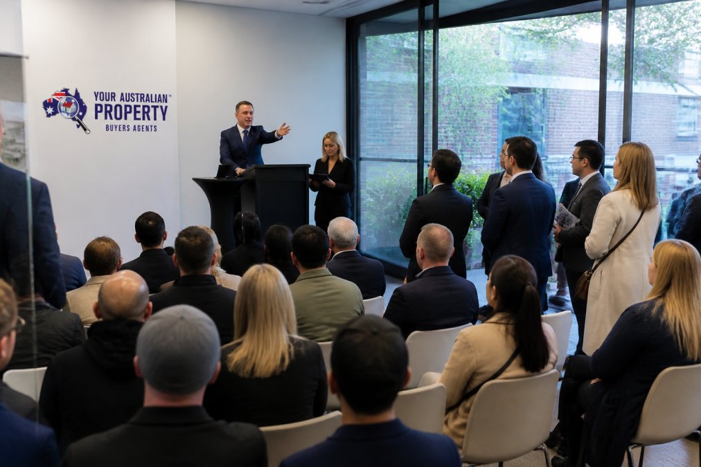 Competitive property auction in progress with auctioneer speaking from podium, bidders seated and standing in modern auction room with natural lighting