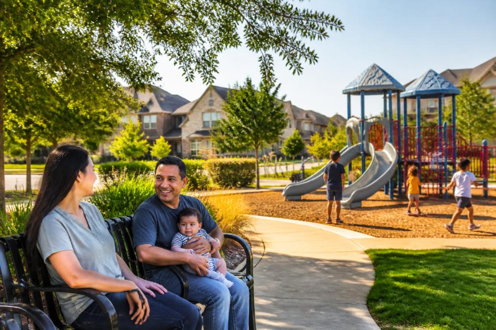 Children playing in a modern community park with colorful playground equipment, families gathered on benches nearby, and well-maintained suburban homes with manicured lawns visible in the distance under bright afternoon sunlight