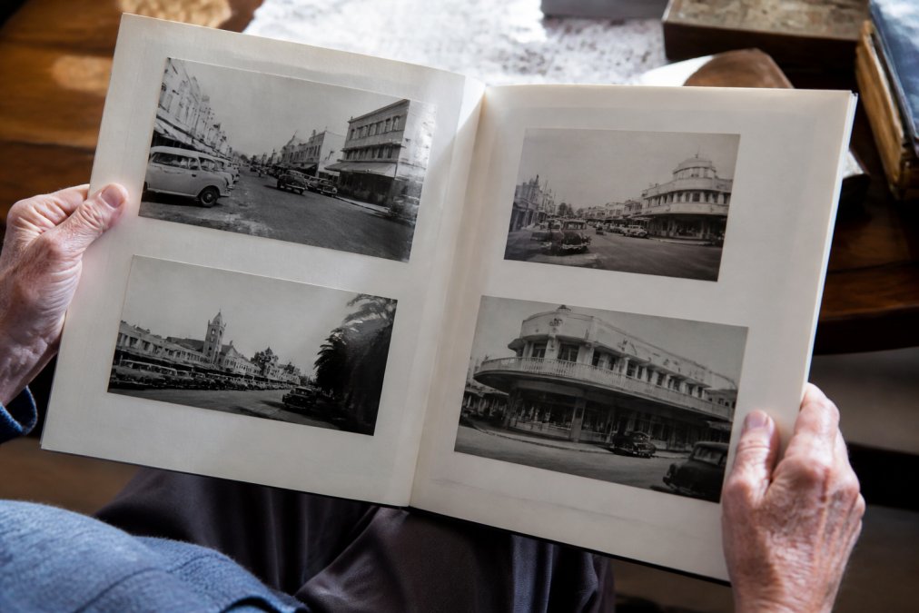 Close-up of weathered hands holding open a vintage photo book with black and white photographs showing Australian street scenes, vintage cars, and period storefronts from the 1950s-1970s, with soft natural light illuminating the pages