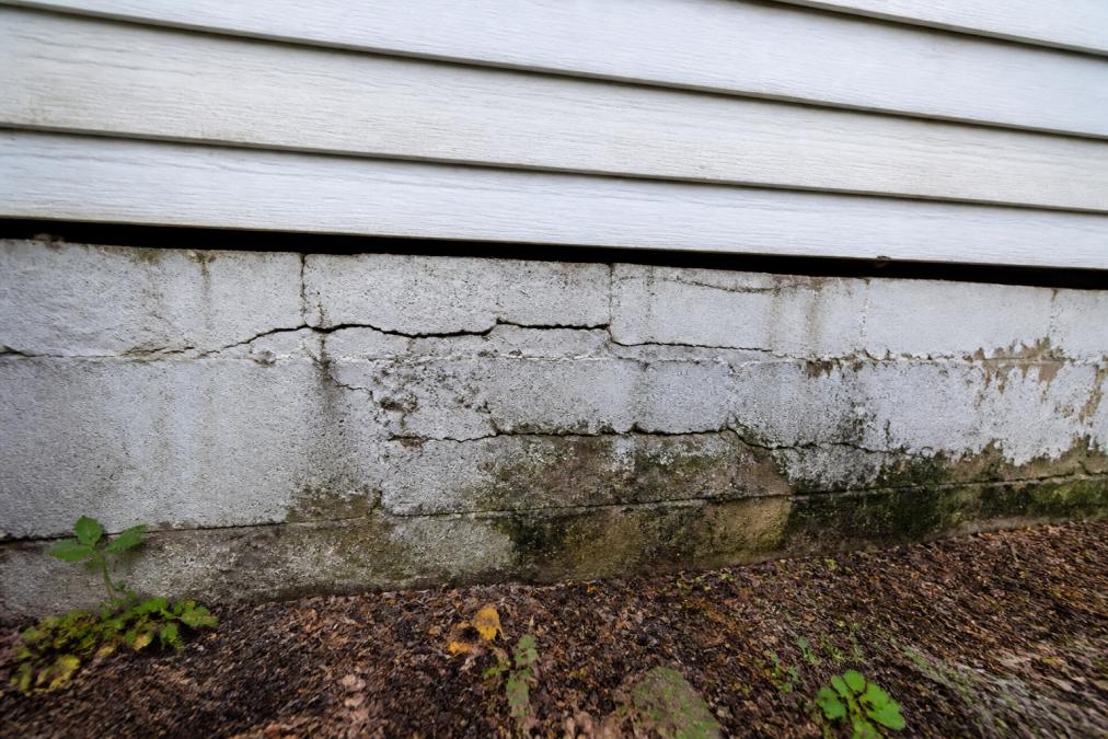 Close-up of a cracked and crumbling concrete foundation wall on an older home, showing visible horizontal cracks and moisture staining, photographed in natural daylight from ground level