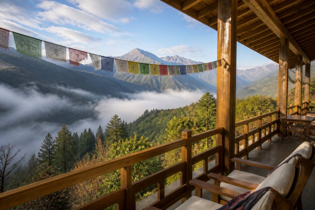 A luxury lodge balcony overlooking a misty Himalayan valley with prayer flags strung across the foreground, morning light catching the mountain ridge