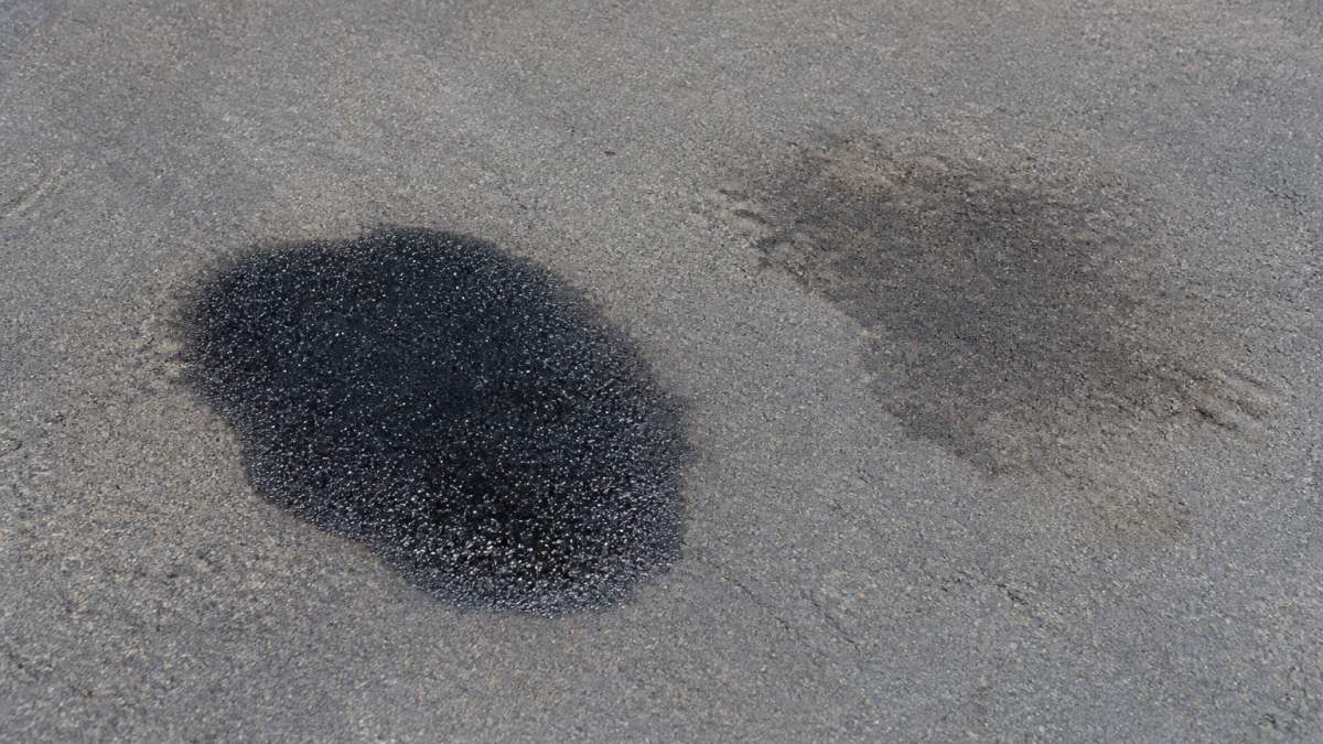 Close-up of fresh dark glossy oil stain next to faded, lighter set-in oil stain on gray asphalt surface in bright daylight