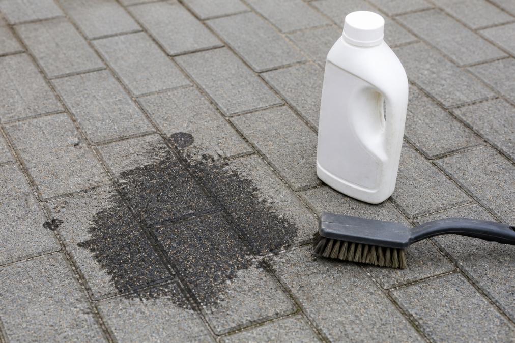 Close-up of fresh dark oil stain on grey block paving with a bottle of degreaser and stiff brush positioned nearby, natural daylight