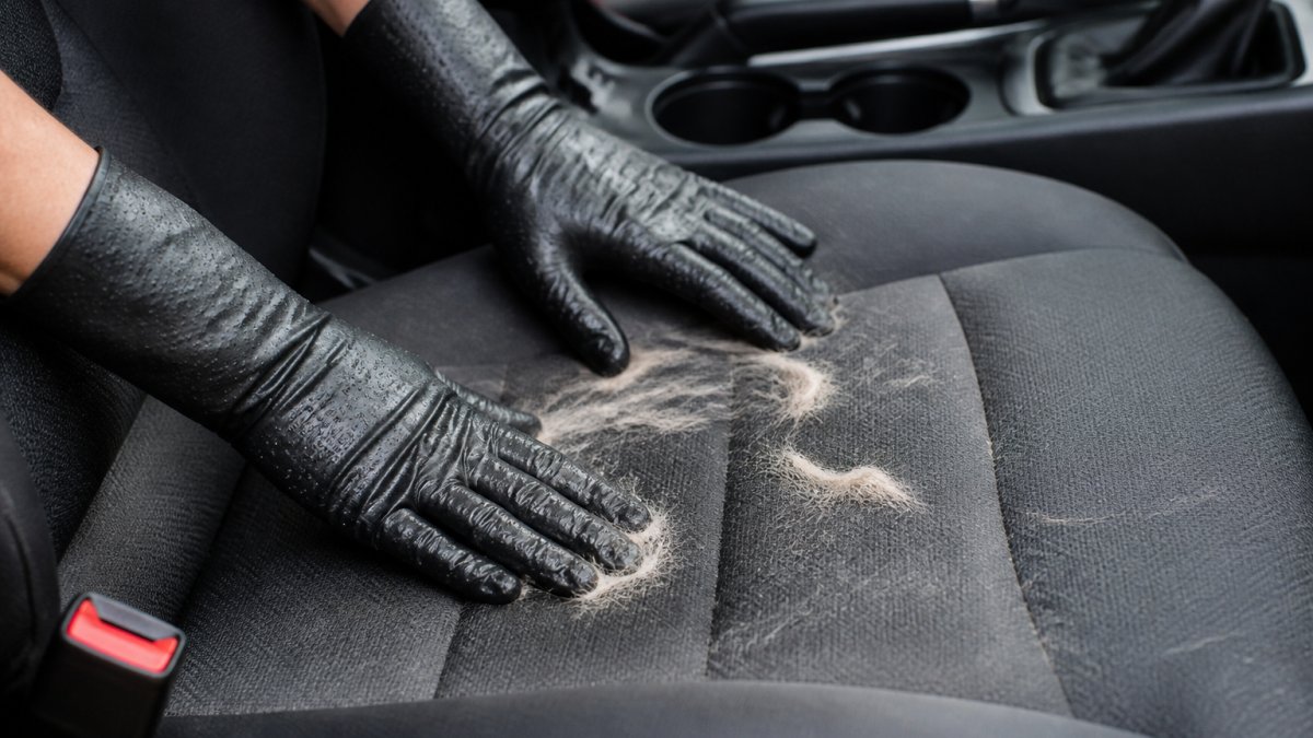 Close-up of hands wearing damp rubber gloves rubbing a fabric car seat, with pet hair visibly lifting and clumping together on the seat surface