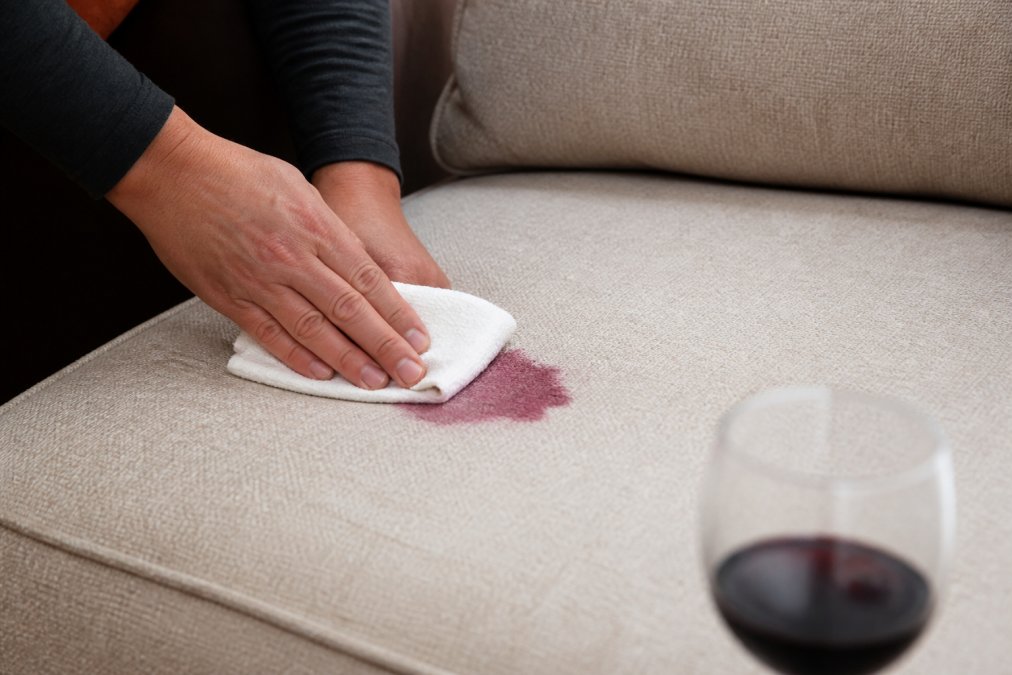 Close-up of hands using a white absorbent cloth to gently blot a fresh red wine stain on beige upholstery, demonstrating proper pressing motion with controlled pressure