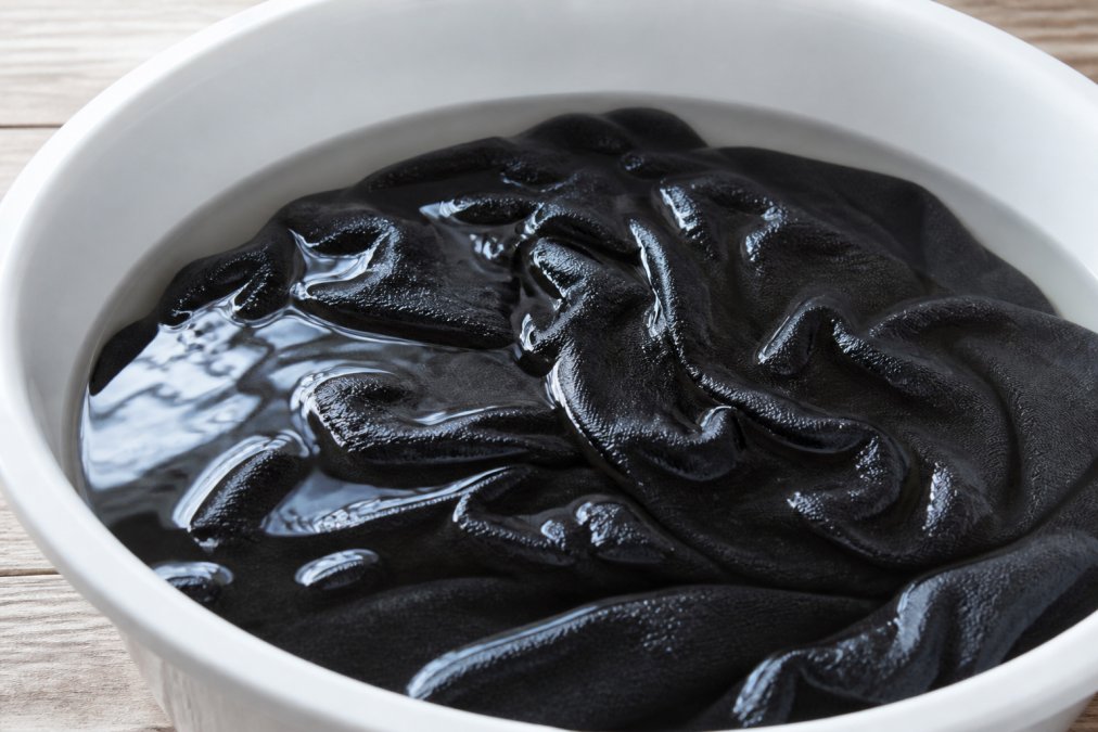 Close-up of dark fabric submerged in clear soak solution with white vinegar in a white ceramic basin, showing fabric fibers absorbing the liquid and light reflecting off the water surface