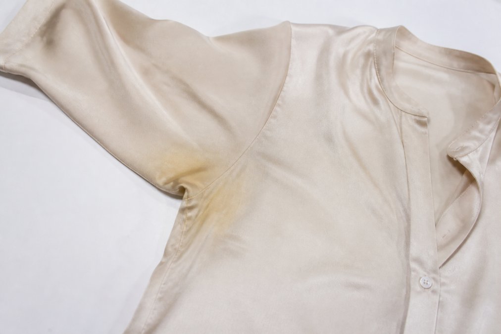 A silk blouse laid flat on a clean white surface with visible perspiration stains under the arms, ready for treatment, showing the characteristic yellowing or discoloration from dried sweat