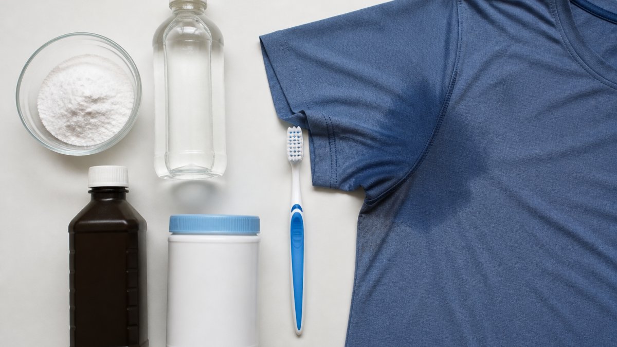 Flat-lay arrangement of stain treatment supplies on a clean white surface: baking soda in a small bowl, white vinegar bottle, hydrogen peroxide bottle, oxygen bleach container, soft-bristled toothbrush, and a folded athletic shirt showing visible sweat stains under the arm