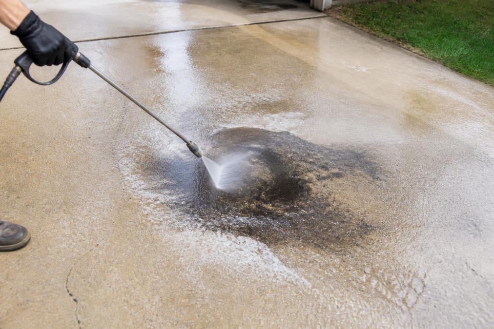 A gloved hand guiding a pressure washer wand over a concrete driveway, with water and suds rinsing away a dark oil stain near the center of the frame