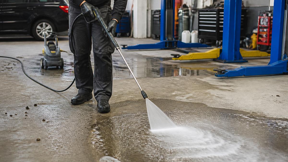 A person in work clothes using a pressure washer with a wide fan nozzle on a concrete workshop floor, water spray visible, tools and a car lift in the background