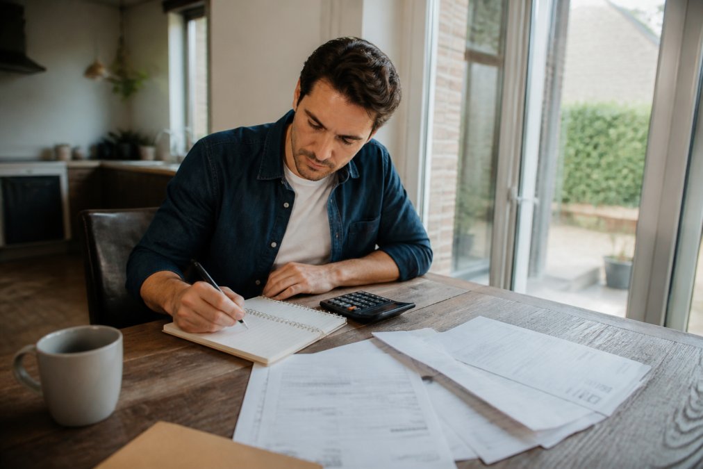Huiseigenaar zittend aan keukentafel met notitieblok, rekenmachine en uitgeprinte offertes, concentrerend op het plannen van renovatiefasen met natuurlijk daglicht