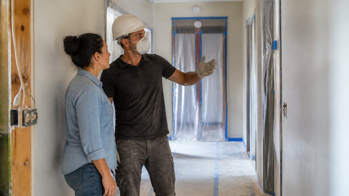A contractor in a hard hat and dust mask walking a tenant through a partially renovated apartment hallway in a four-unit building, with protective floor coverings and sealed doorways visible behind them