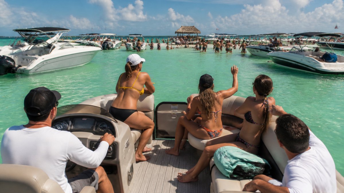 A group of friends on a pontoon boat approaching a crowded sandbar in Miami, with clear turquoise water and other boats moored nearby under a sunny sky