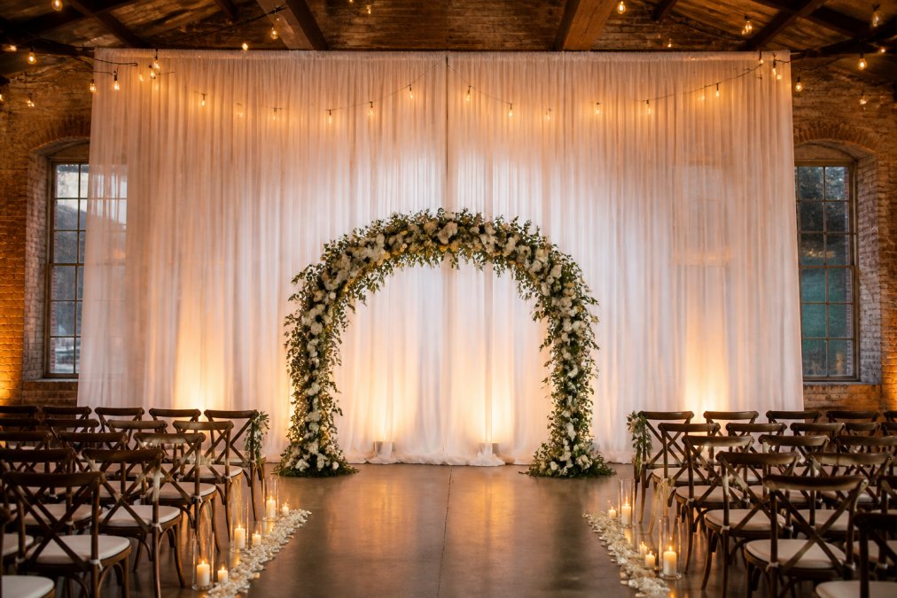 An elegant wedding ceremony backdrop featuring floor-to-ceiling white sheer voile draping with soft warm ambient lighting behind a floral arch in a converted warehouse venue with exposed brick walls and Edison bulb string lights overhead