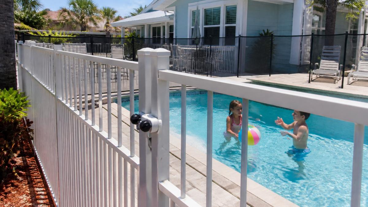 A family with two young children playing in a fenced private pool at a beach cottage, with a self-latching gate visible and pool safety fence in the background