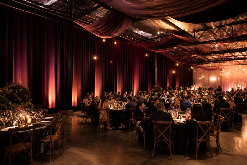 Warehouse wedding reception transformed with elegant floor-to-ceiling velvet drapes in deep jewel tones, uplighting casting warm amber light on the fabric, industrial ceiling partially hidden by ceiling draping, guests seated at tables with the dramatic backdrop visible