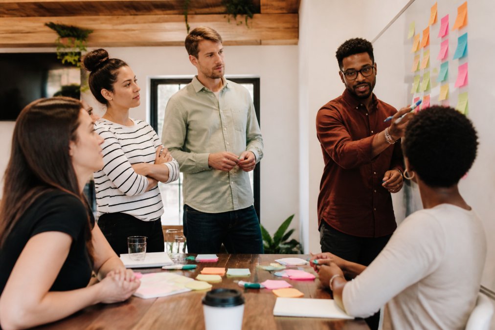 Diverse group of professionals collaborating around a whiteboard during a strategy session, with sticky notes and colorful markers visible on the board and table