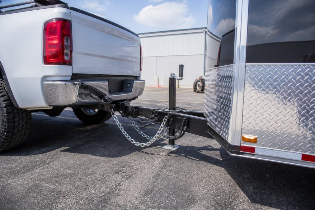 A heavy-duty pickup truck hitched to a fully loaded enclosed car hauler trailer in a parking lot, showing the trailer hitch, safety chains, and electric brake connection visible under afternoon sunlight