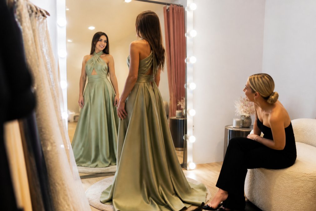A woman adjusting a flowing designer dress in a boutique mirror while a friend offers style advice