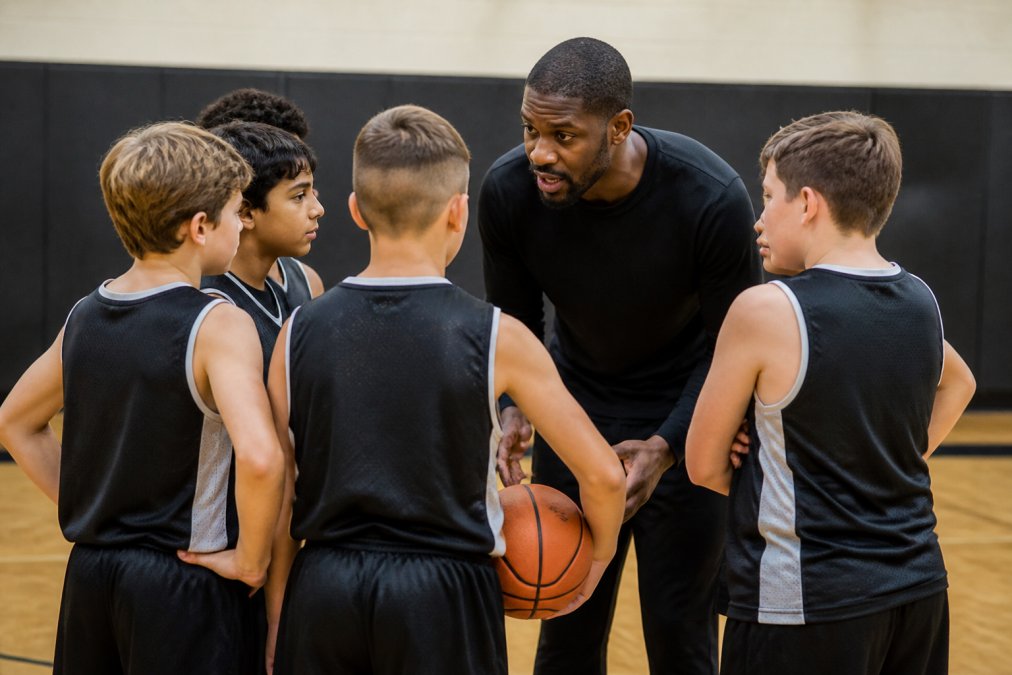 Young basketball players gathered in a team huddle with coach providing instruction, showing focused engagement and teamwork during practice session with natural gym lighting