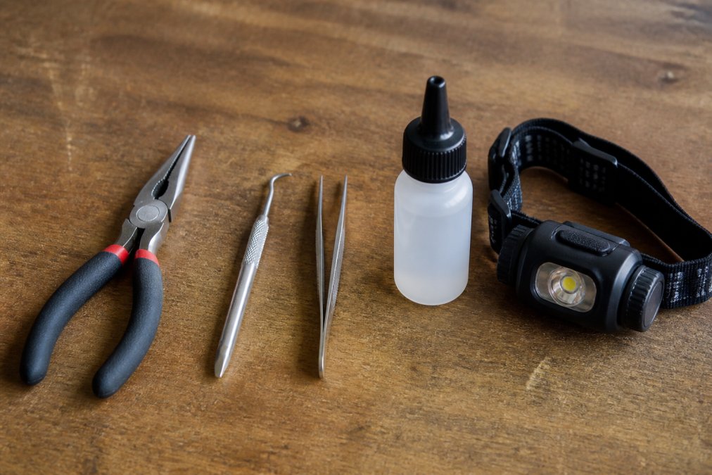 Close-up of a workbench displaying key extraction tools including needle-nose pliers with smooth grip, metal key extractor pick with hook, tweezers, small bottle of silicone spray lubricant, and LED headlamp arranged in organized rows on a clean surface