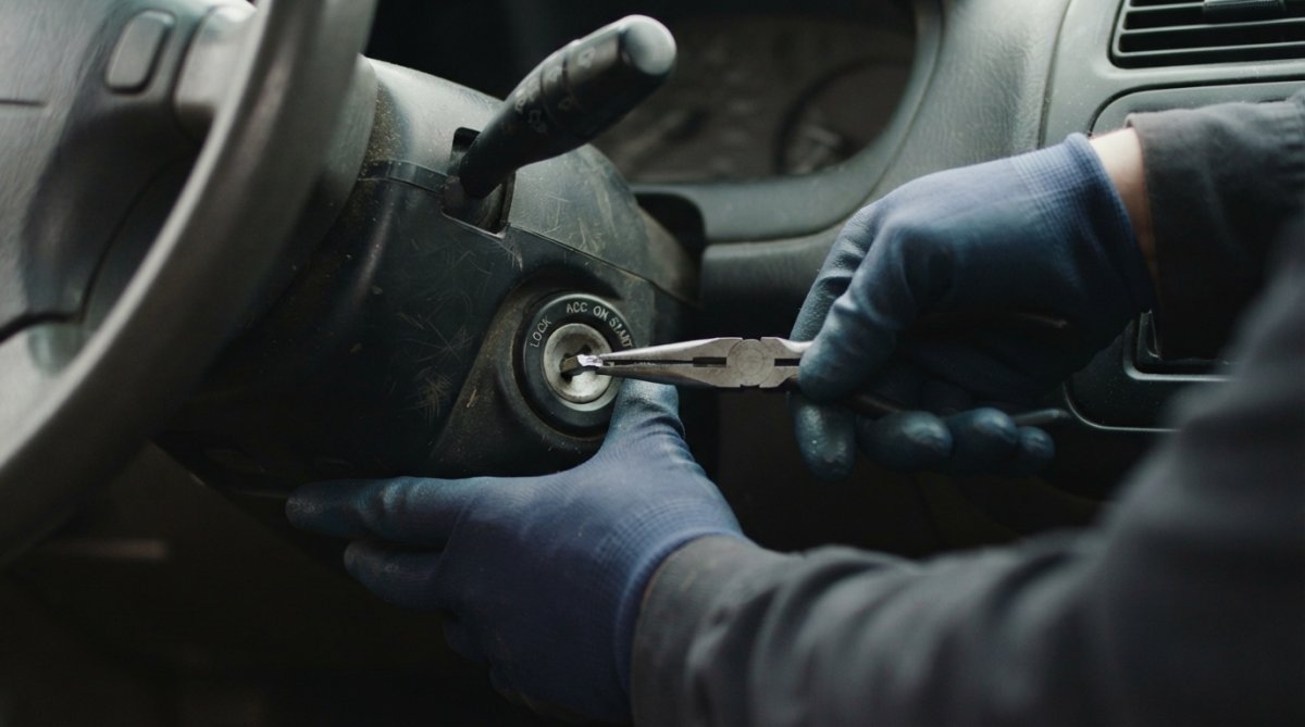 Hands wearing work gloves carefully using needle-nose pliers to extract a broken key fragment from a car ignition cylinder, showing proper grip angle and straight pulling technique with natural workshop lighting