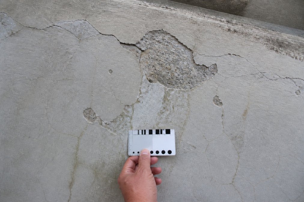 Close-up of concrete balcony surface showing visible spalling, hairline cracks, and water staining patterns indicating structural degradation, with professional measuring gauge for scale