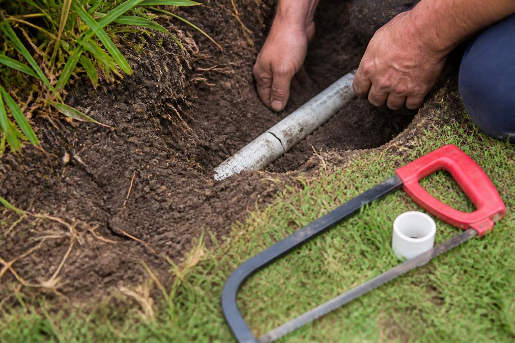 Close-up of a technician's hands carefully excavating around a white PVC irrigation pipe in a garden bed, exposing a visible hairline crack, with a hacksaw and slip-fix coupling laid out on the grass beside the shallow trench, in bright afternoon sunlight