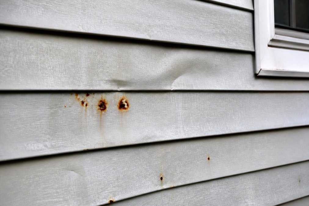 Close-up of weathered aluminum siding on older residential home showing visible dents, rust-colored corrosion spots, and chalky faded paint texture in natural daylight