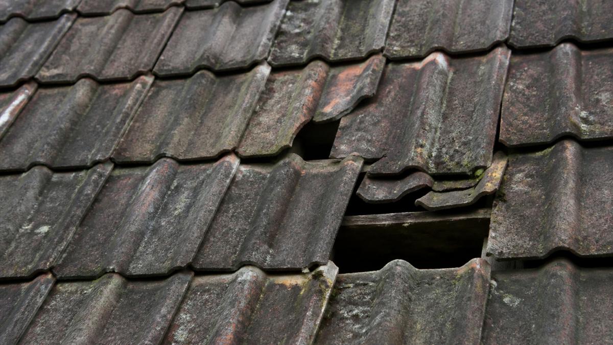 Close-up of weathered roof tiles showing cracked, curled, and missing sections with visible wear patterns and water staining on the surface