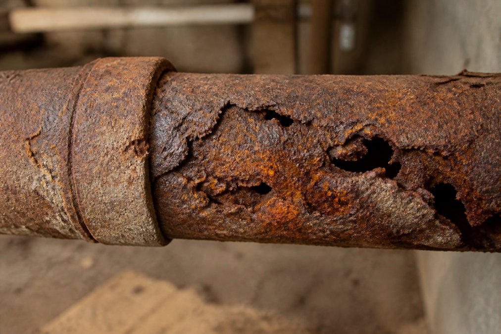Close-up of heavily corroded cast iron pipe showing deep rust pitting, orange oxidation stains, and brittle sections with visible structural deterioration and mineral scale buildup