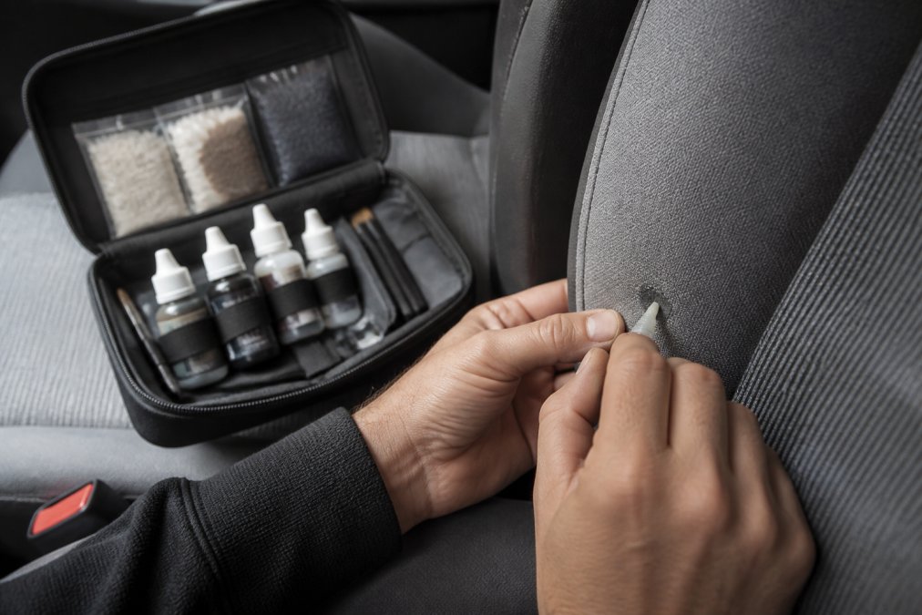 Close-up of hands using a small applicator tool to carefully apply colour pigment to a repaired section of grey car seat fabric, with a repair kit open nearby showing bottles of adhesive and synthetic fibers
