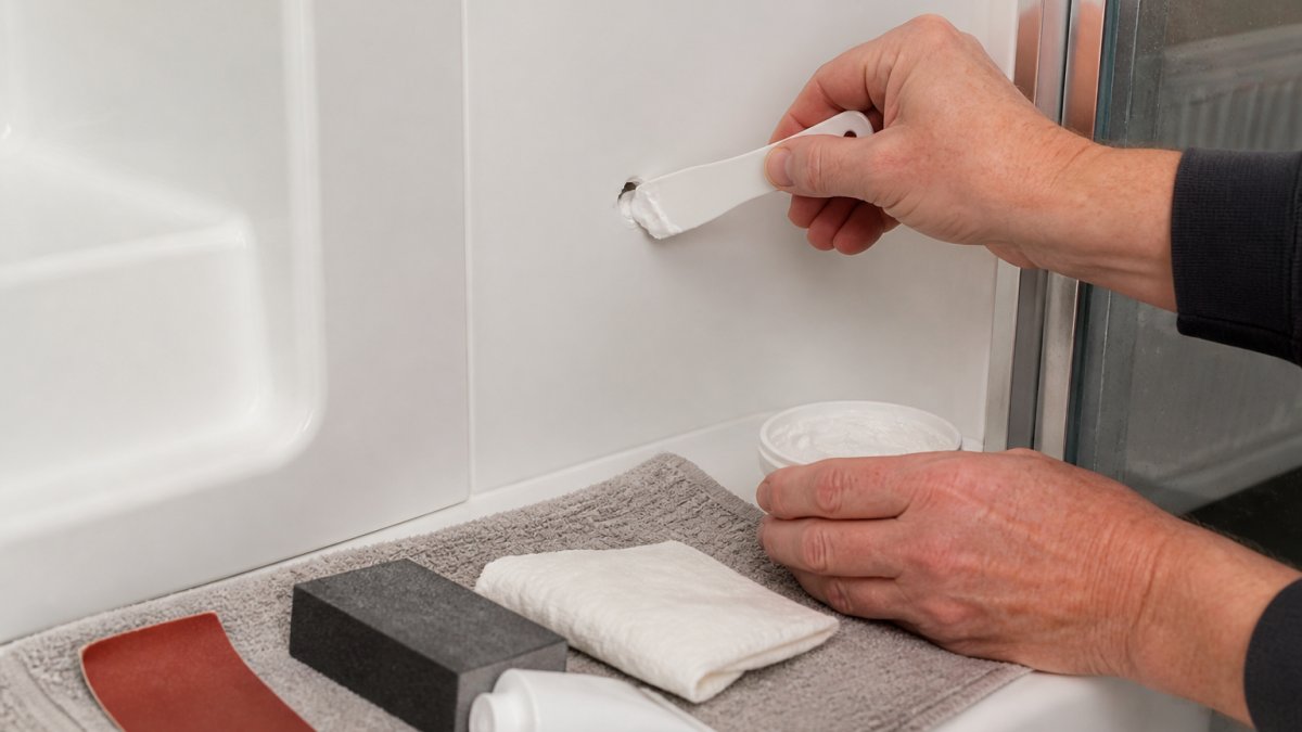Close-up of a homeowner's hands applying white filler to a small hole in a PVC shower wall panel with a plastic spatula, in a bright bathroom