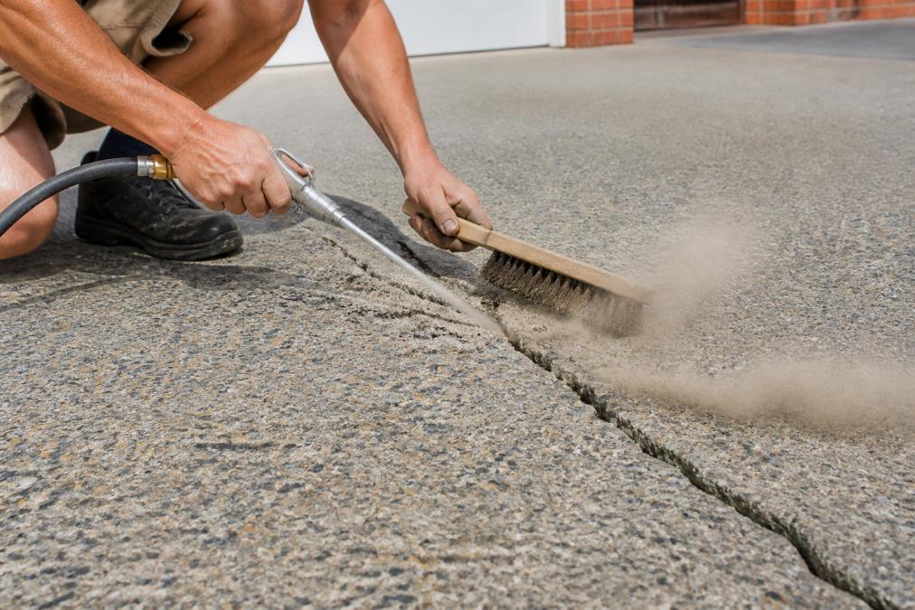 Professional using compressed air and wire brush to clean deep debris from a concrete crack in spray-on driveway surface, showing dust particles being blown out
