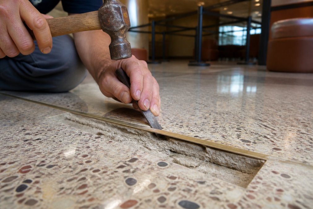 Professional technician's hands using a small chisel and hammer to carefully inspect and remove a loose brass terrazzo divider strip, revealing the substrate and old adhesive residue underneath in a well-lit commercial setting