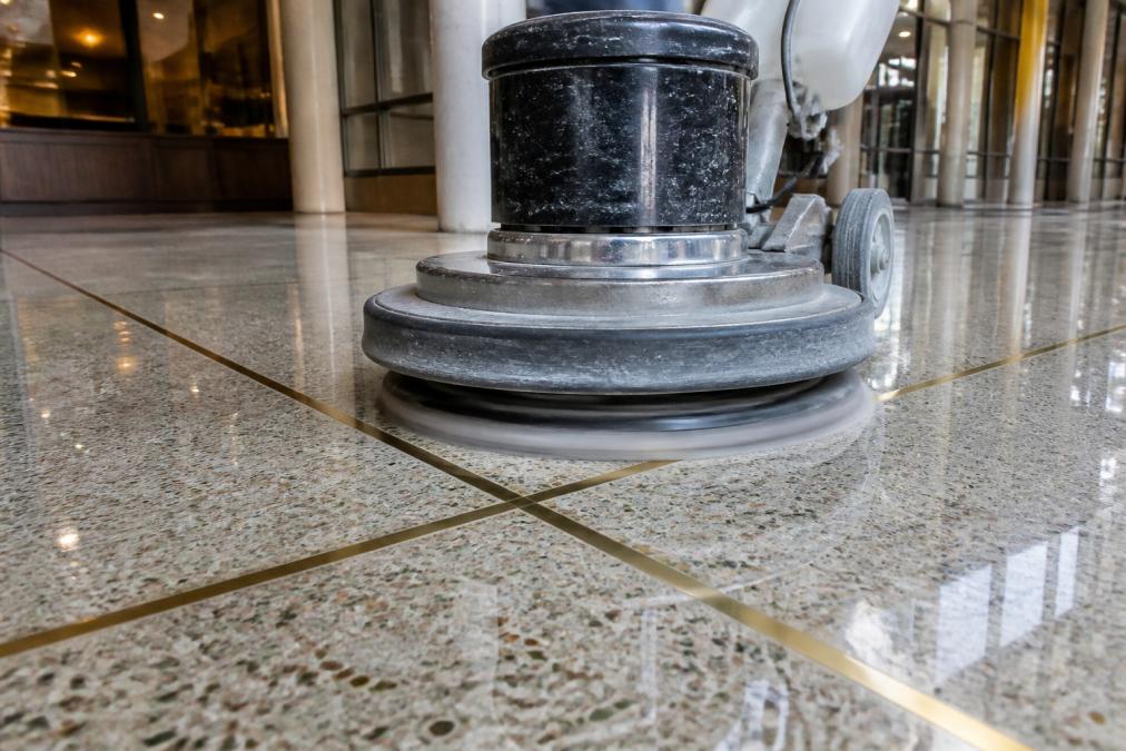 Commercial-grade terrazzo polishing machine with rotating diamond pads actively polishing a terrazzo floor with brass divider strips, showing the restored aggregate exposure and reflective shine under bright facility lighting