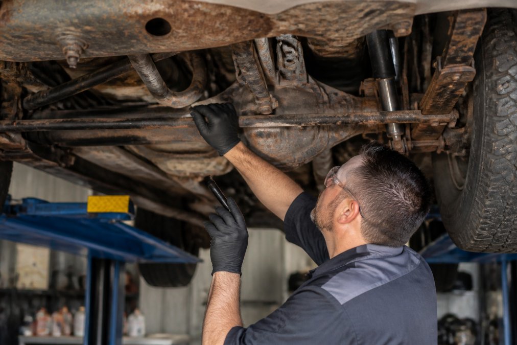 Technician inspecting undercarriage of older truck on lift, checking suspension components and frame for rust and wear, close-up of inspection points and corrosion patterns
