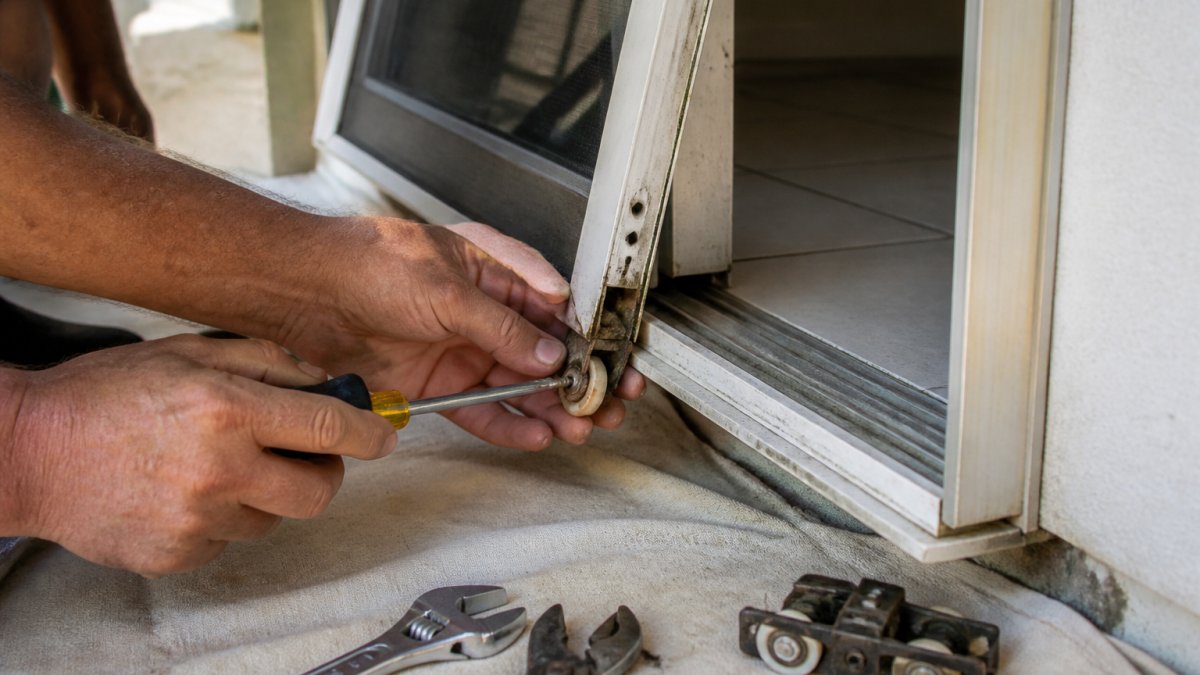 Hands using a screwdriver to remove a worn roller while repairing sliding glass doors vs replacing the unit.