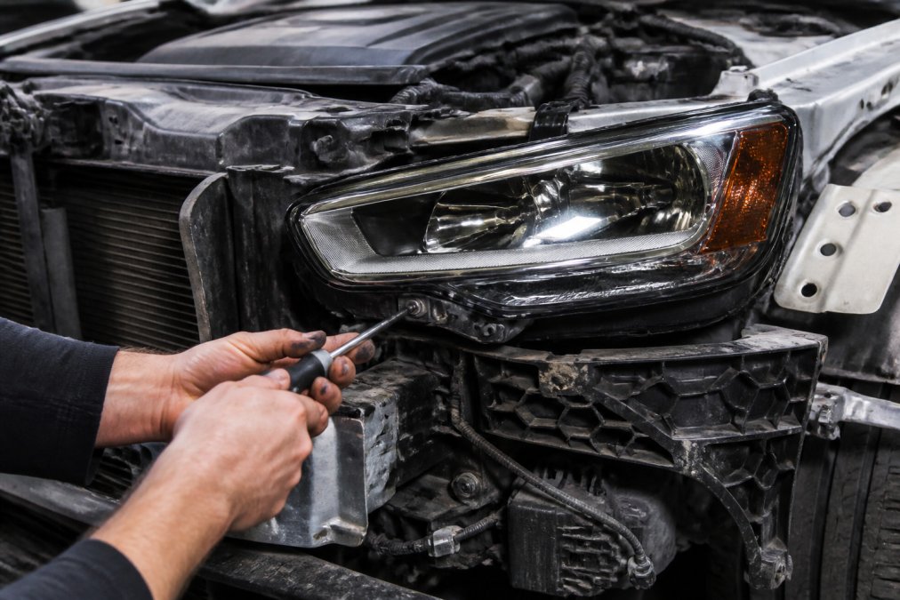 Technician's hands using a Torx T25 screwdriver to remove bolts from an Audi A4 B9 headlight assembly mounting bracket, with the front bumper already removed and the connector visible in the background