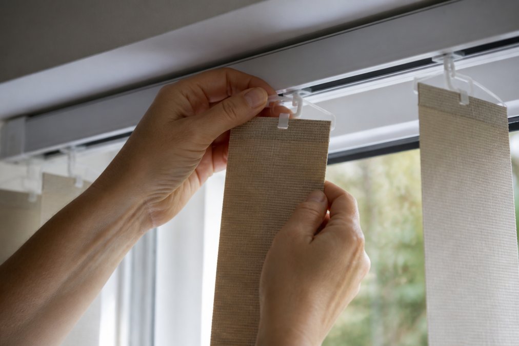 Hands carefully releasing a vertical blind slat from its top hanger, showing the connection point between the slat carrier hook and the headrail track, with natural window light