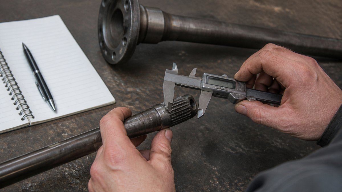 Close-up of a mechanic's hands using digital calipers to measure the splined end of a removed axle shaft on a workbench, with a notebook and a second shaft in the background