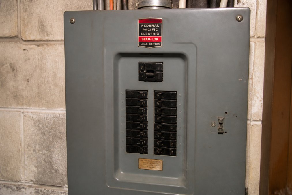 Close-up of a Federal Pacific Stab-Lok electrical panel mounted on a basement wall, showing distinctive breaker design and "Federal Pacific Electric" labeling clearly visible