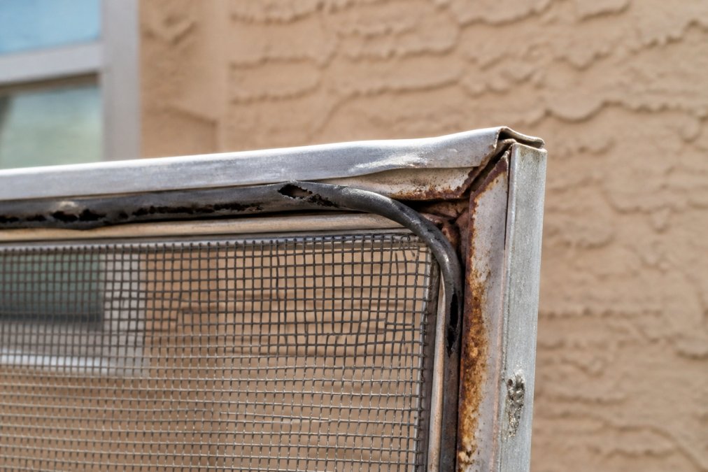Close-up of corroded window screen frame corner showing rust buildup, bent aluminum edges, and deteriorated spline rubber with visible sun damage in bright Arizona sunlight