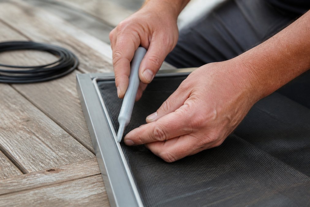 Hands using a plastic spline tool to press new screen mesh firmly into the aluminum window frame groove, showing proper technique with the spline rubber being seated during professional rescreening installation