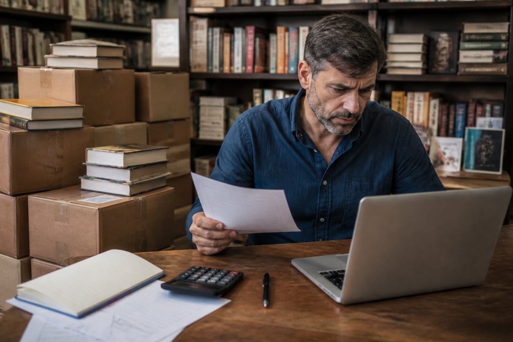 Libraio gestisce manualmente i resi distributori editoriali tra pile di libri e fogli di calcolo in negozio