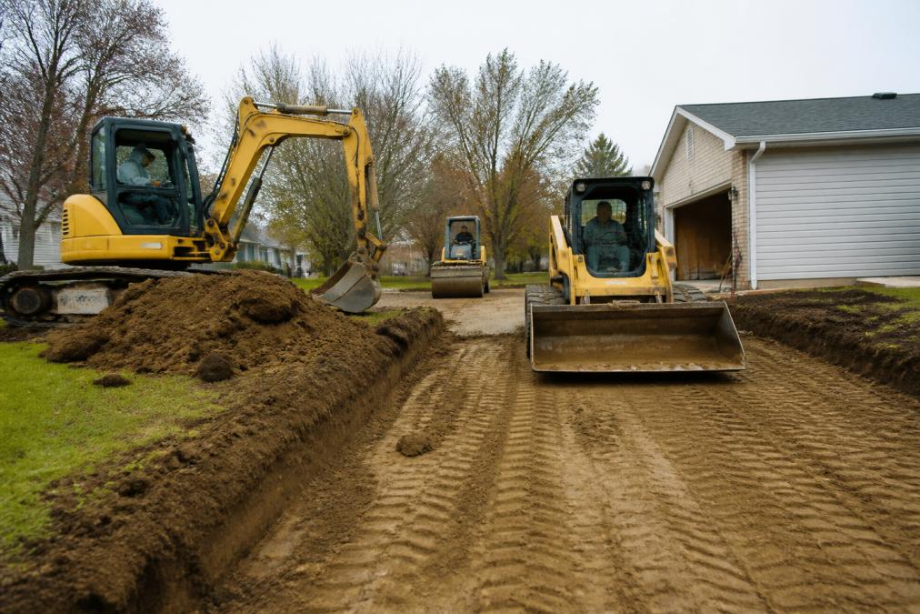 Excavation crew operating heavy machinery on residential property, moving dirt and grading the driveway base, with compacted soil visible and equipment positioned for base preparation