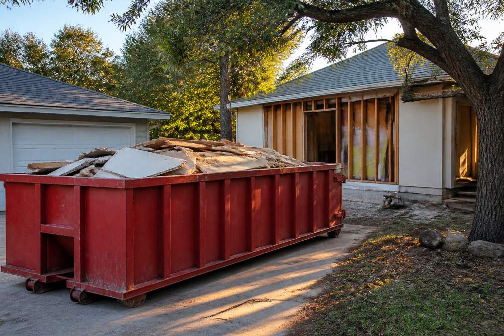 A red roll-off dumpster parked on a residential concrete driveway next to a home under renovation, with visible construction debris including drywall and lumber inside the container, late afternoon sunlight casting shadows across the yard